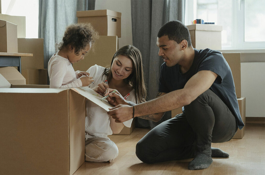 Father and stepmother with child writing on a box during a move, representing how after divorce you can struggle with stepparenting but work together to feel happier as a blended family.